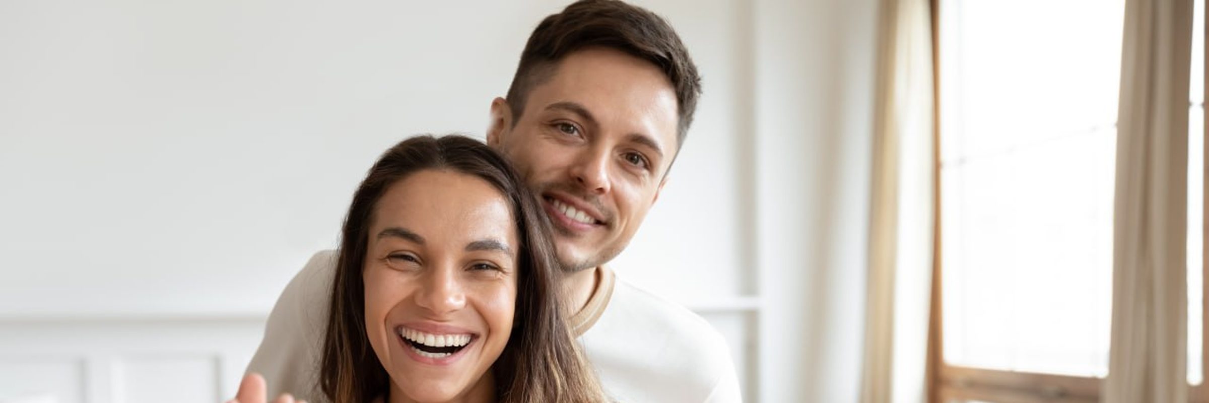 A couple holds up the keys to their new home, laughing together in a bright living room.
