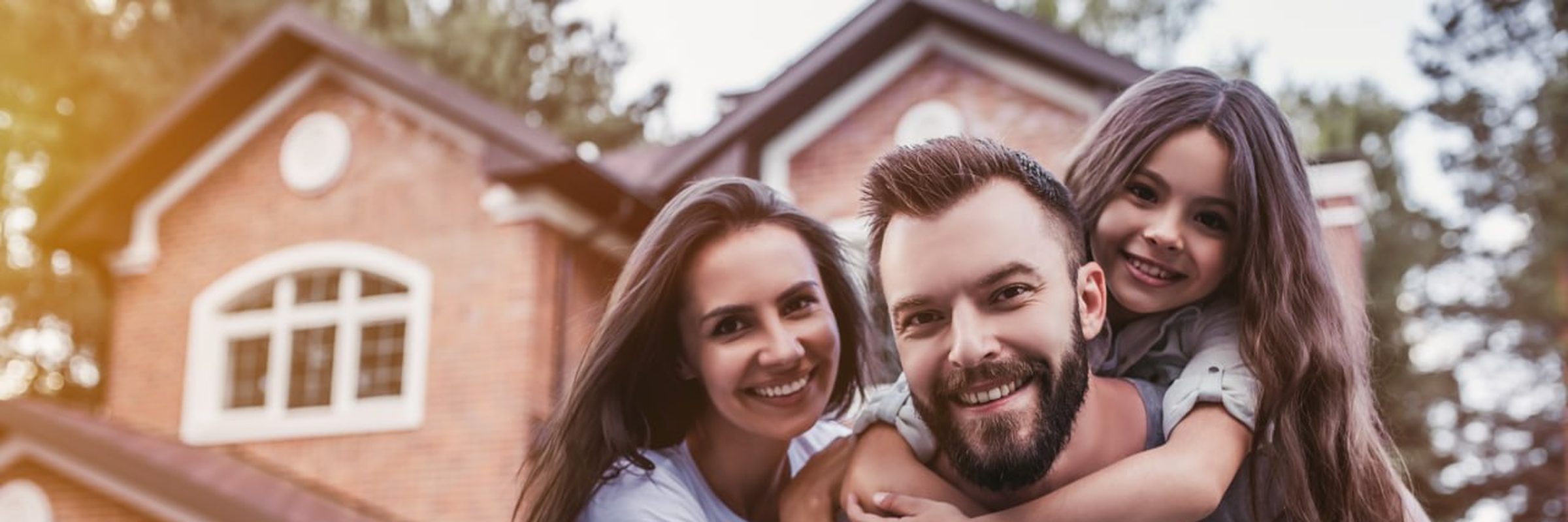A family of three smiles together in front of their brick suburban home during golden hour.